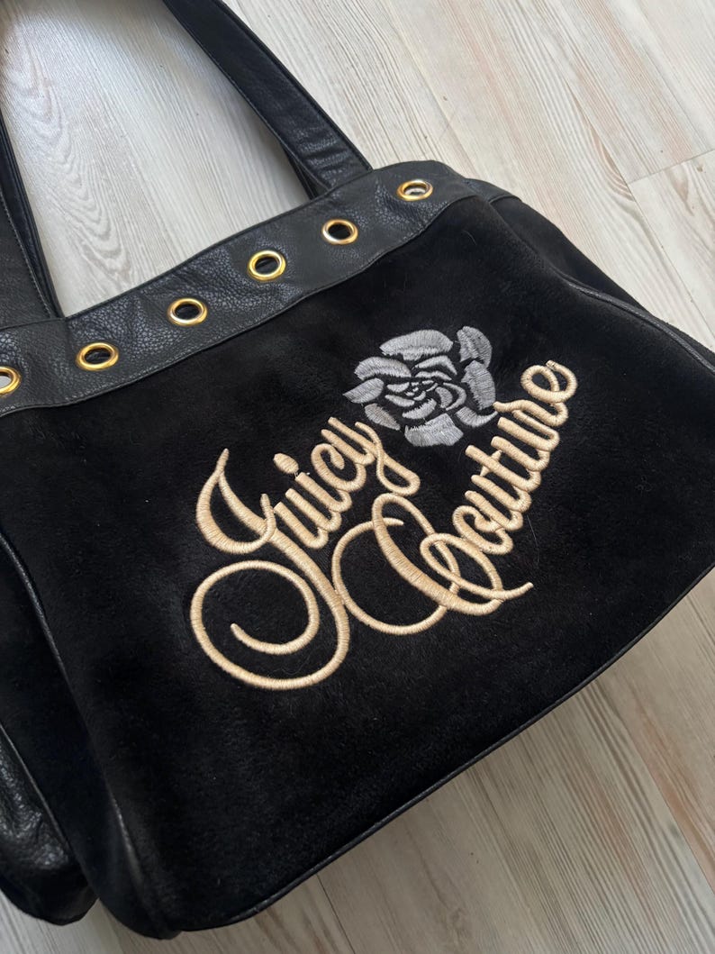 May include: Black tote bag with the embroidered text "Juicy Couture" and a rose graphic. The bag features black leather straps and gold-colored grommets along the top edge. The bag is made of a soft, velvety material.