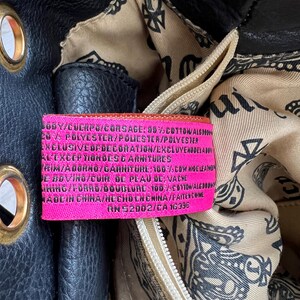 May include: Close-up of a black leather handbag with gold-colored grommets. The interior lining features a paisley pattern. A pink tag with white text details the bag's composition and origin.
