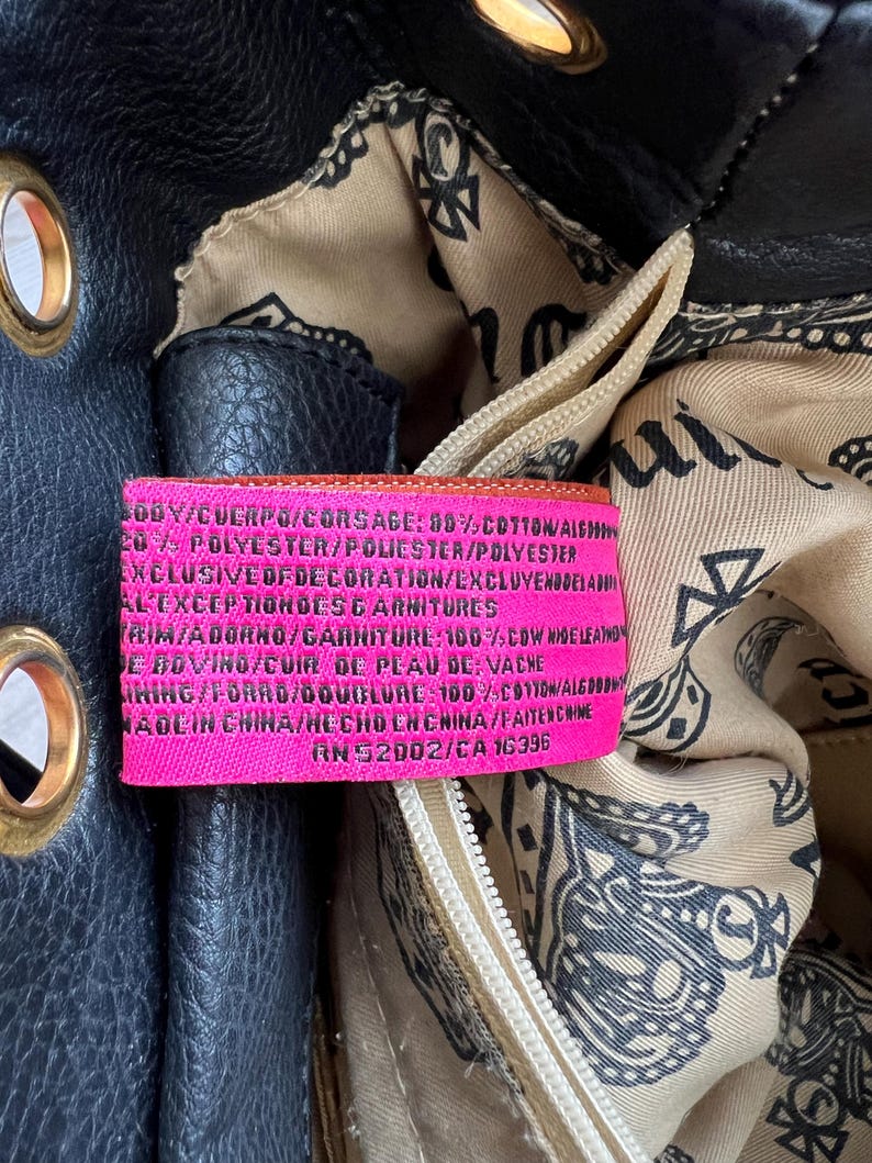 May include: Close-up of a black leather handbag with gold-colored grommets. The interior lining features a paisley pattern. A pink tag with white text details the bag's composition and origin.