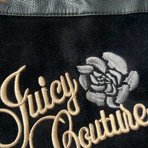 May include: Close-up of a black fabric with the embroidered text "Juicy Couture" in a gold script font. A gray rose is embroidered next to the text. The top edge features black leather with gold grommets.
