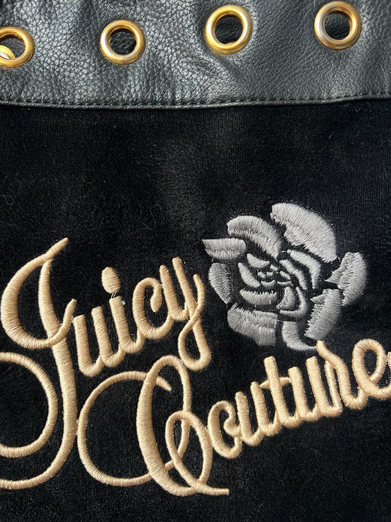 May include: Close-up of a black fabric with the embroidered text "Juicy Couture" in a gold script font. A gray rose is embroidered next to the text. The top edge features black leather with gold grommets.