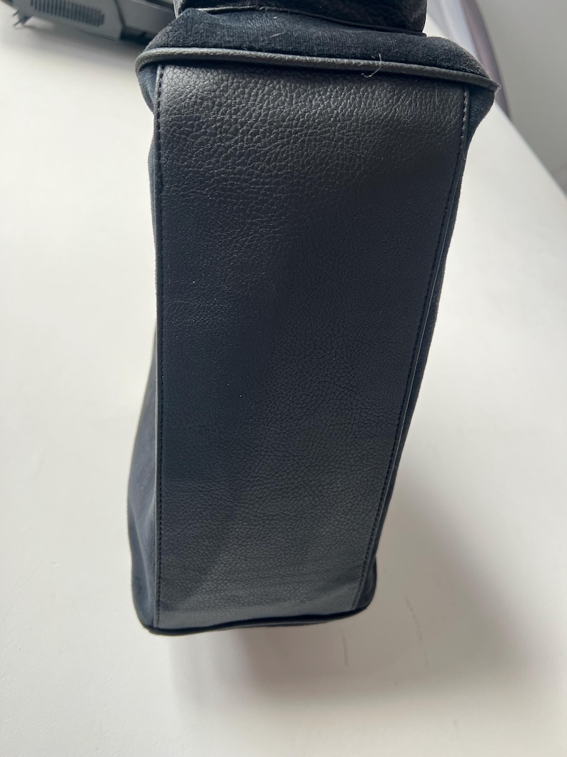 May include: A black handbag with a textured leather exterior and a suede top. The bag has a rectangular shape and appears to be empty. The stitching is visible along the seams.