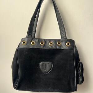May include: Black tote bag with a suede body and leather trim. Features gold-tone grommets along the top edge and a small side pocket. The bag has a leather handle and a heart-shaped emblem with the letter "J".