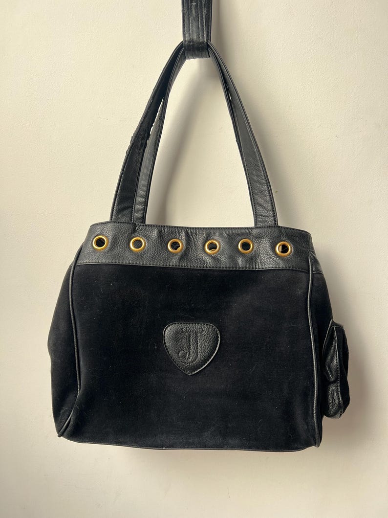 May include: Black tote bag with a suede body and leather trim. Features gold-tone grommets along the top edge and a small side pocket. The bag has a leather handle and a heart-shaped emblem with the letter "J".