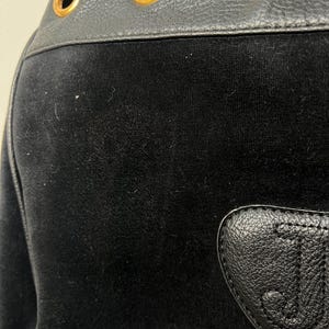 May include: Close-up of a black velour garment with a leather accent. The leather trim features three gold-colored grommets and a heart-shaped patch with the initials "J.C." embossed on it.