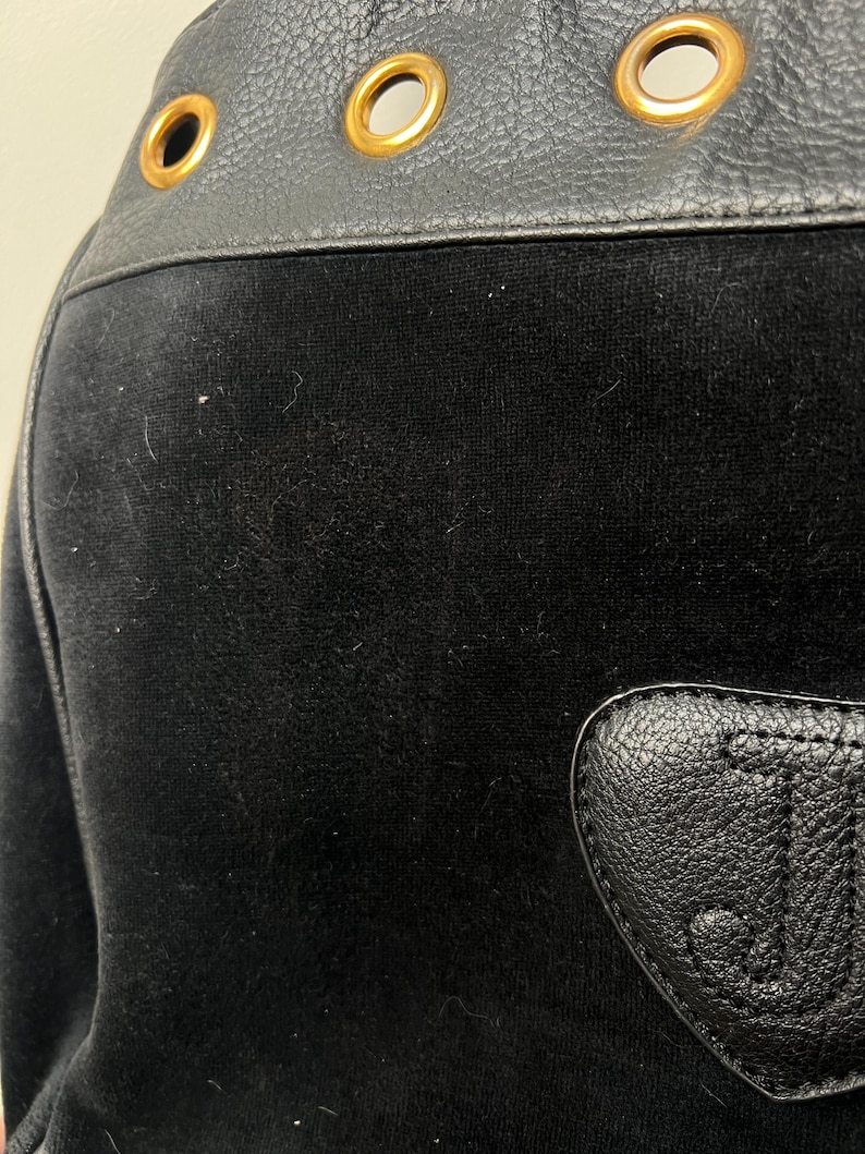 May include: Close-up of a black velour garment with a leather accent. The leather trim features three gold-colored grommets and a heart-shaped patch with the initials "J.C." embossed on it.