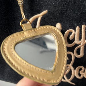 May include: A gold-colored heart-shaped mirror with a matching strap and clasp. The mirror is attached to a black fabric with gold embroidery. The mirror is a small, handheld accessory.