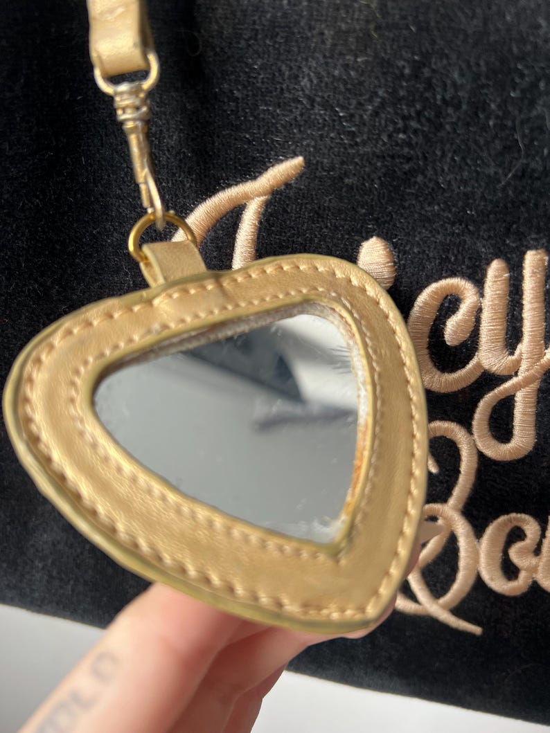 May include: A gold-colored heart-shaped mirror with a matching strap and clasp. The mirror is attached to a black fabric with gold embroidery. The mirror is a small, handheld accessory.