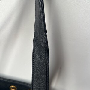 May include: Close-up of a black leather handbag strap. The strap shows signs of wear and tear, with visible scuffs and damage. The bag's body is partially visible, featuring gold-colored hardware.