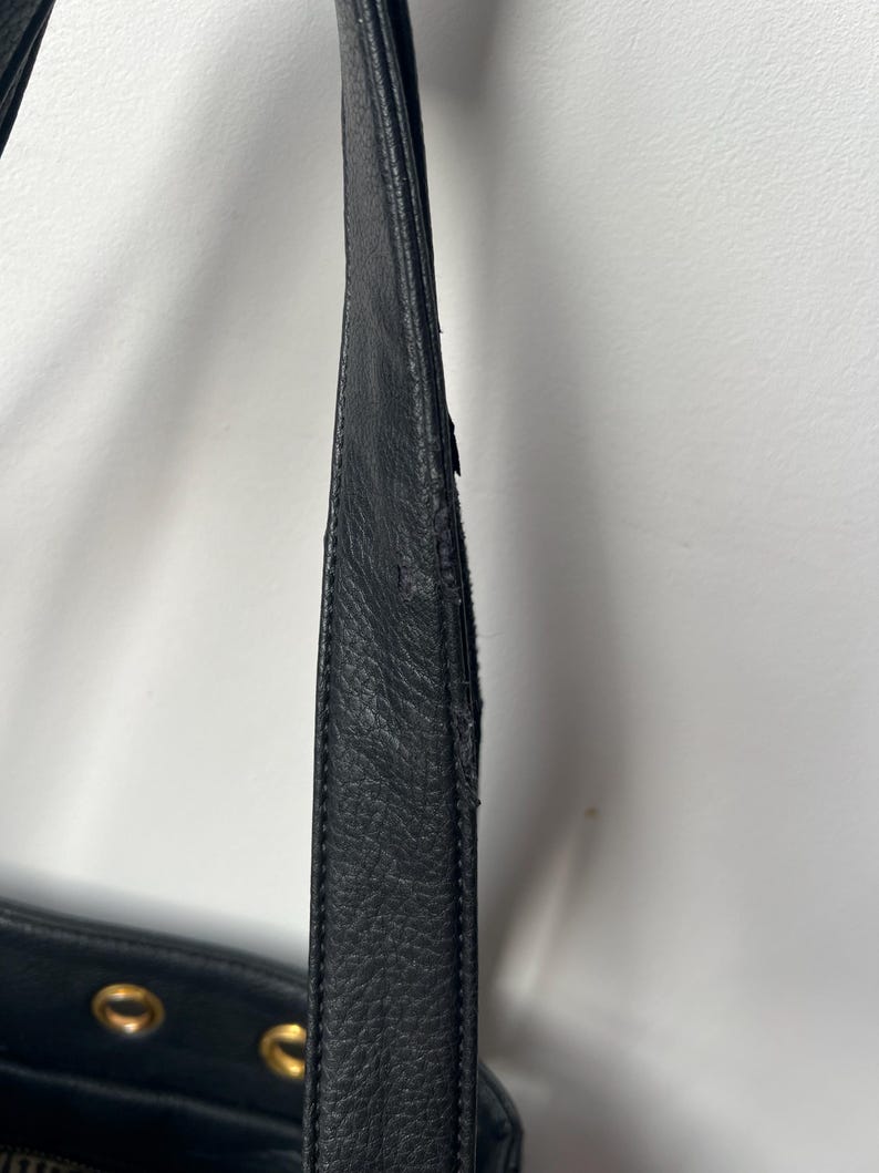 May include: Close-up of a black leather handbag strap. The strap shows signs of wear and tear, with visible scuffs and damage. The bag's body is partially visible, featuring gold-colored hardware.