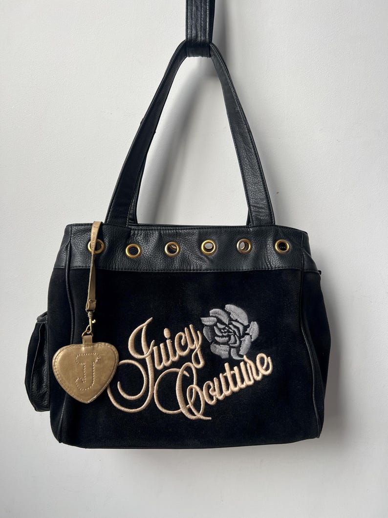 May include: Black suede tote bag with leather handles and trim. Features the embroidered text "Juicy Couture" with a rose design. Includes a gold heart-shaped charm with the letter "J".