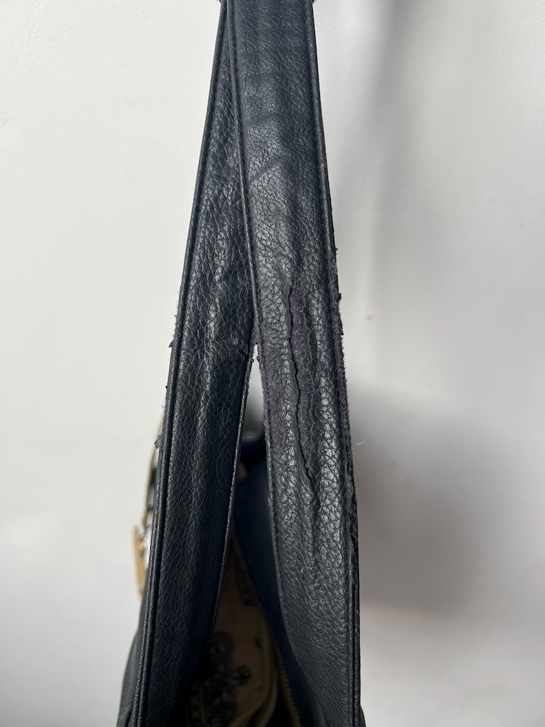 May include: Black leather shoulder bag with a textured surface. The bag's strap shows signs of wear and tear. The interior of the bag is visible, revealing a patterned fabric lining. The bag is hanging against a white background.