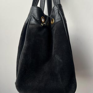 May include: Black suede tote bag with leather accents and handles. The bag features gold-colored grommets at the top and a drawstring closure. The bag is designed with a rounded bottom.