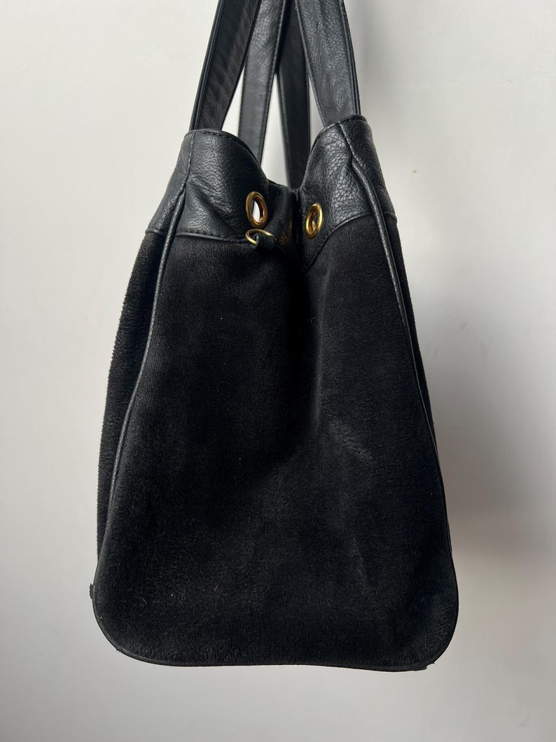 May include: Black suede tote bag with leather accents and handles. The bag features gold-colored grommets at the top and a drawstring closure. The bag is designed with a rounded bottom.