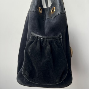 May include: Black handbag with a suede-like texture and leather accents. The bag features a drawstring closure, a front pocket, and gold-colored hardware. The handles are made of black leather.