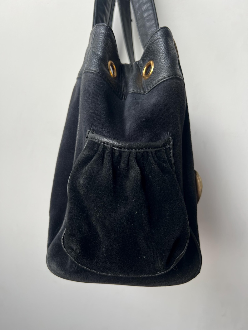 May include: Black handbag with a suede-like texture and leather accents. The bag features a drawstring closure, a front pocket, and gold-colored hardware. The handles are made of black leather.
