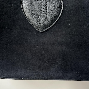 May include: Close-up of a black bag with a heart-shaped leather patch. The patch features the letter "J" in a cursive font. The bag has a soft, velvety texture.