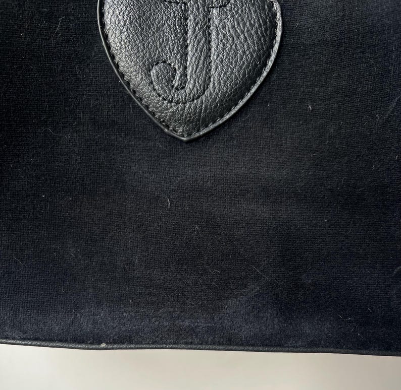 May include: Close-up of a black bag with a heart-shaped leather patch. The patch features the letter "J" in a cursive font. The bag has a soft, velvety texture.