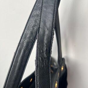 May include: Close-up of a black handbag with visible wear and tear on the strap. The bag features gold-colored rivets and a textured leather-like material. The strap shows signs of peeling and damage.