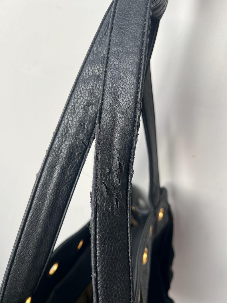 May include: Close-up of a black handbag with visible wear and tear on the strap. The bag features gold-colored rivets and a textured leather-like material. The strap shows signs of peeling and damage.