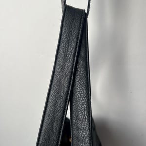 May include: Close-up of a black leather shoulder bag with textured straps. The bag is hanging against a white background, showcasing the details of the material and design. The straps are thick and appear durable.