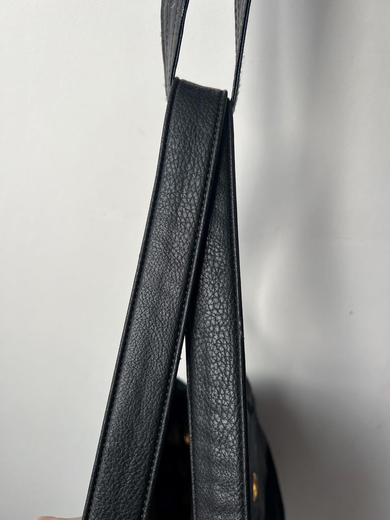 May include: Close-up of a black leather shoulder bag with textured straps. The bag is hanging against a white background, showcasing the details of the material and design. The straps are thick and appear durable.