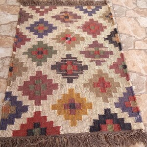 Op de afbeelding: Rechthoekig jute vloerkleed met een geometrisch patroon in tinten rood, paars, groen en geel. Het vloerkleed heeft een beige achtergrond en een gefranjerde rand. Het patroon bestaat uit ruitvormen.