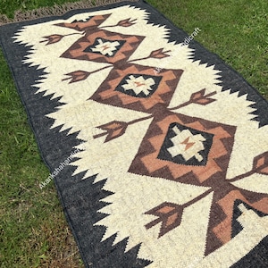 Puede incluir: Alfombra rectangular con un campo central crema con patrones geométricos marrones y negros. La alfombra tiene un borde gris oscuro con un borde en zigzag y flecos marrones.