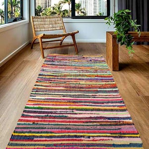 Könnte beinhalten: Ein bunter, rechteckiger Teppich mit horizontalen Streifen in verschiedenen Farben, darunter Rosa, Gelb, Blau und Grün. Der Teppich liegt auf einem Holzboden, mit einem Holzstuhl und einem Fenster im Hintergrund.