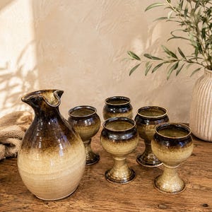 May include: A ceramic sake set consisting of one rounded carafe with a flared spout and four matching stemmed cups. Each piece features a two-tone glaze with a dark brown finish at the top that fades into a light beige colour at the base.