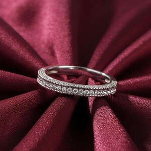 May include: A silver ring with a double row of small, clear gemstones. The ring is set against a deep red satin fabric, creating a contrast of textures and colors. The gemstones sparkle, reflecting light.