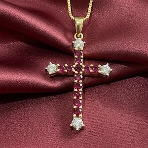 Pode incluir: Um colar com pingente de cruz dourada com uma corrente delicada. A cruz é adornada com pequenas pedras preciosas vermelhas redondas e quatro pedras maiores, claras, em forma de diamante. O pingente repousa sobre um tecido vermelho escuro, semelhante ao cetim.