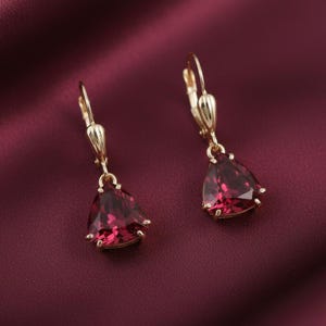 May include: A pair of gold-colored drop earrings with triangular, deep red gemstones. The earrings have a lever-back closure and are set against a burgundy background.