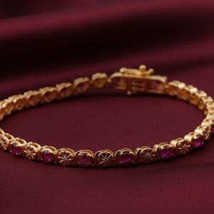 Op de afbeelding: Een goudkleurige armband met ovale, rode edelstenen gezet in een bloemmotief. De armband ligt op een donkerrode achtergrond en heeft een sluiting.