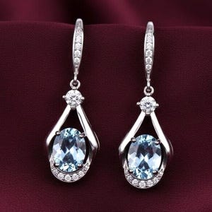 Puede incluir: Un par de pendientes colgantes plateados con piedras preciosas ovaladas de color azul claro. Los pendientes presentan una pequeña gema redonda y transparente en la parte superior y una media luna de gemas transparentes más pequeñas en la parte inferior.