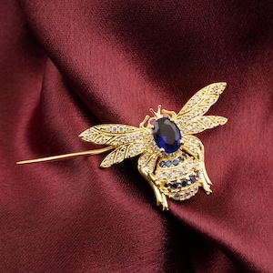 Puede incluir: Broche de abeja dorada con una gran gema azul ovalada en el centro. Las alas están detalladas con pequeñas piedras transparentes. El cuerpo de la abeja presenta piedras azules y transparentes. El broche está sobre una tela roja oscura.