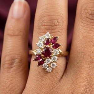 May include: A gold ring featuring a cluster of round and marquise-cut rubies and clear white diamonds. The stones are arranged in an asymmetrical, elongated floral-inspired pattern set on a polished gold band that encircles the finger.