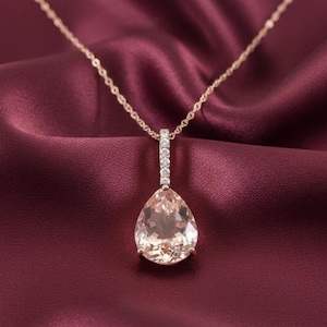 Puede incluir: Un collar de oro rosa con un colgante en forma de pera. El colgante es de color melocotón claro y está adornado con una fila de pequeñas piedras transparentes. La delicada cadena dorada contrasta con un fondo burdeos.
