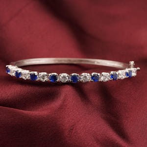 Könnte beinhalten: Ein silbernes Armreif-Armband mit abwechselnd runden blauen und klaren Edelsteinen. Das Armband liegt auf einem tiefroten Hintergrund, der das Funkeln der Steine hervorhebt.