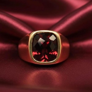 Peut inclure: Une bague dorée ornée d'une grosse pierre précieuse rouge foncé taillée en carré. La pierre est facettée et sertie dans un sertissage en or. La bague est présentée sur un tissu de satin rouge riche, créant un effet luxueux.