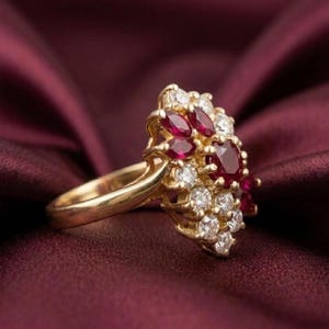May include: A gold ring featuring a cluster of marquise-cut red rubies and round-cut white diamonds. The stones are arranged in a dense, multi-layered floral pattern set upon a polished gold band, showcasing a vibrant contrast between the red and white.