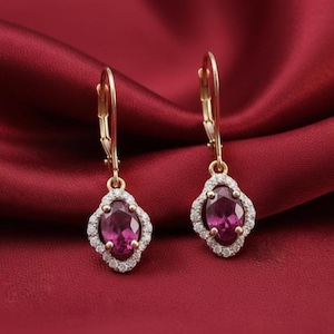 May include: A pair of gold-coloured drop earrings. Each earring features a large, oval, magenta-coloured gemstone surrounded by a halo of small, clear gemstones. The earrings hang from a gold-coloured lever back.