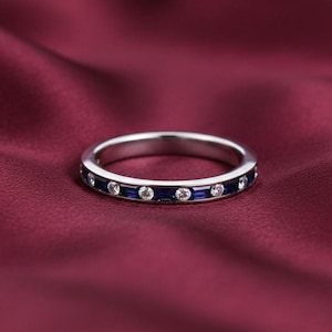 May include: A silver ring with alternating blue and clear gemstones. The ring is set against a deep red background, highlighting the sparkle of the stones. The ring's design is simple and elegant, suitable for everyday wear or special occasions.