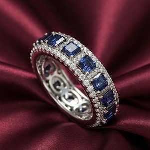 May include: A silver ring with a continuous row of square, deep blue gemstones, interspersed with smaller, round, clear stones. The ring is set against a deep red, textured background, highlighting the sparkle of the gems.