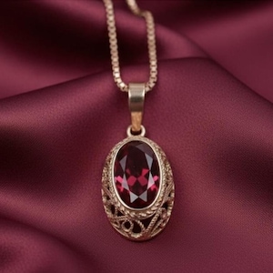 Puede incluir: Un collar con colgante dorado con una piedra preciosa ovalada de color rojo intenso. La piedra preciosa está engastada en un marco dorado calado y decorativo. La cadena del collar es una fina cadena de eslabones dorados, exhibida sobre tela burdeos.