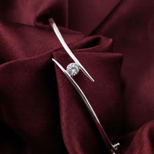May include: A silver-toned bracelet with a single, clear, round gemstone. The bracelet has a modern design, with two curved, parallel bars that cradle the gemstone. The background is a deep burgundy fabric.