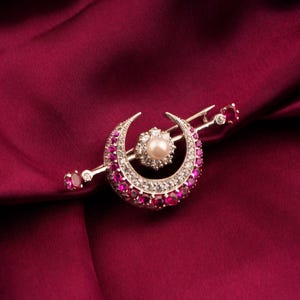 May include: A crescent moon brooch with a pearl center and a halo of white gemstones. The moon's curve is lined with red gemstones. A straight bar extends from the moon, with a red gemstone at each end. The brooch is set against a deep red satin background.