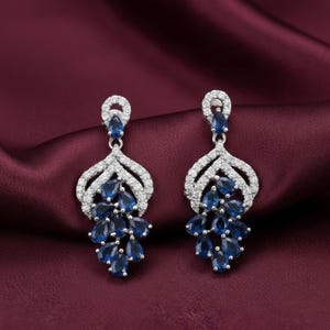 Könnte beinhalten: Ein Paar elegante Ohrhänger mit silberfarbenem Metalldesign. Die Ohrringe sind mit funkelnden, klaren Steinen und tropfenförmigen blauen Edelsteinen besetzt. Der Hintergrund ist ein tiefes Burgunderrot.