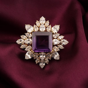 May include: A gold-toned brooch with a square, deep purple center stone. The brooch is surrounded by numerous clear, teardrop-shaped stones, creating a starburst design. The background is a deep burgundy fabric.