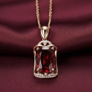 May include: A gold pendant necklace with a large, rectangular, deep red gemstone. The pendant is set with smaller, clear gemstones and hangs from a delicate chain. The necklace is shown against a rich, burgundy fabric.